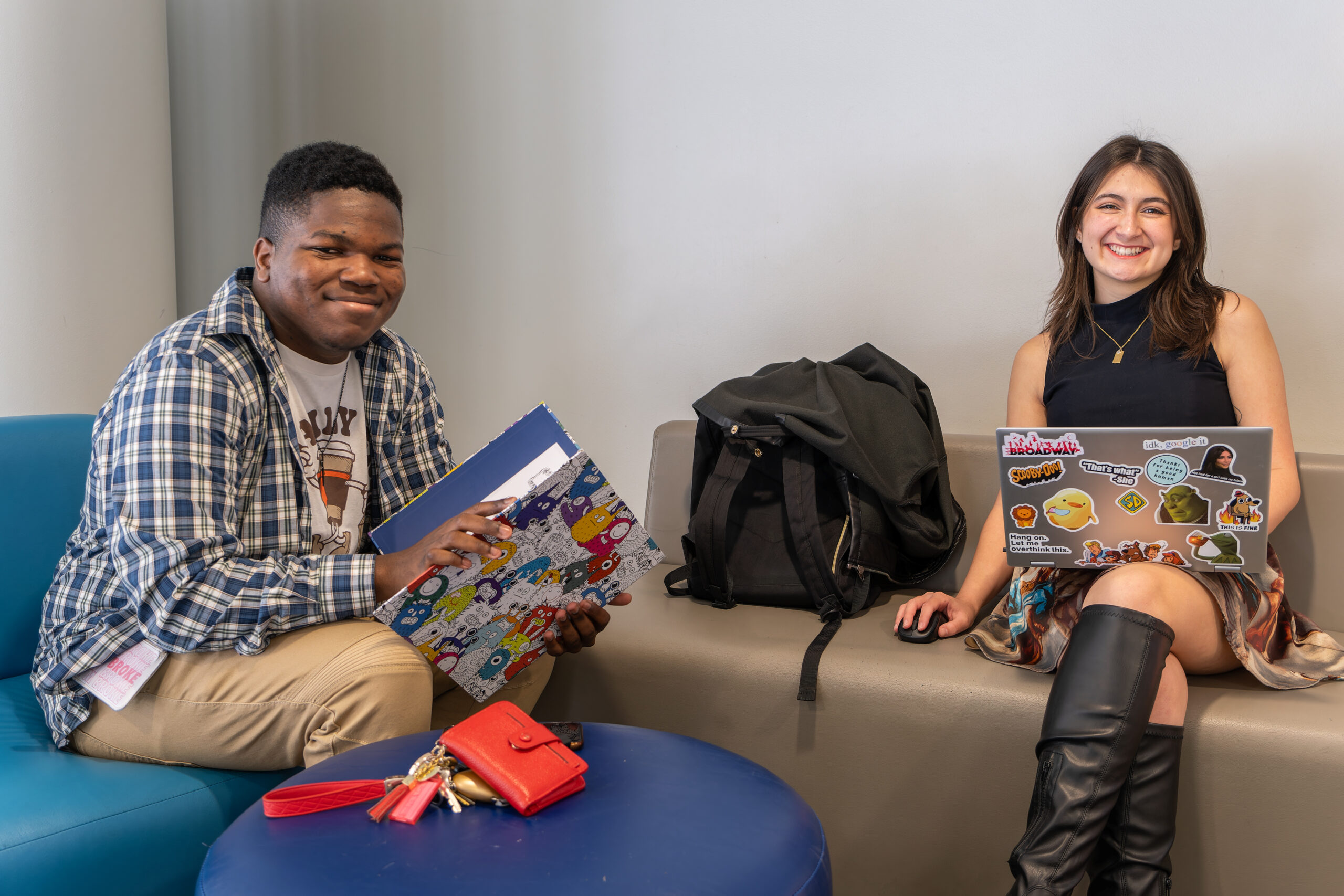 Two UNCG students sit on couches with a book, laptop, wallet, and backpack.