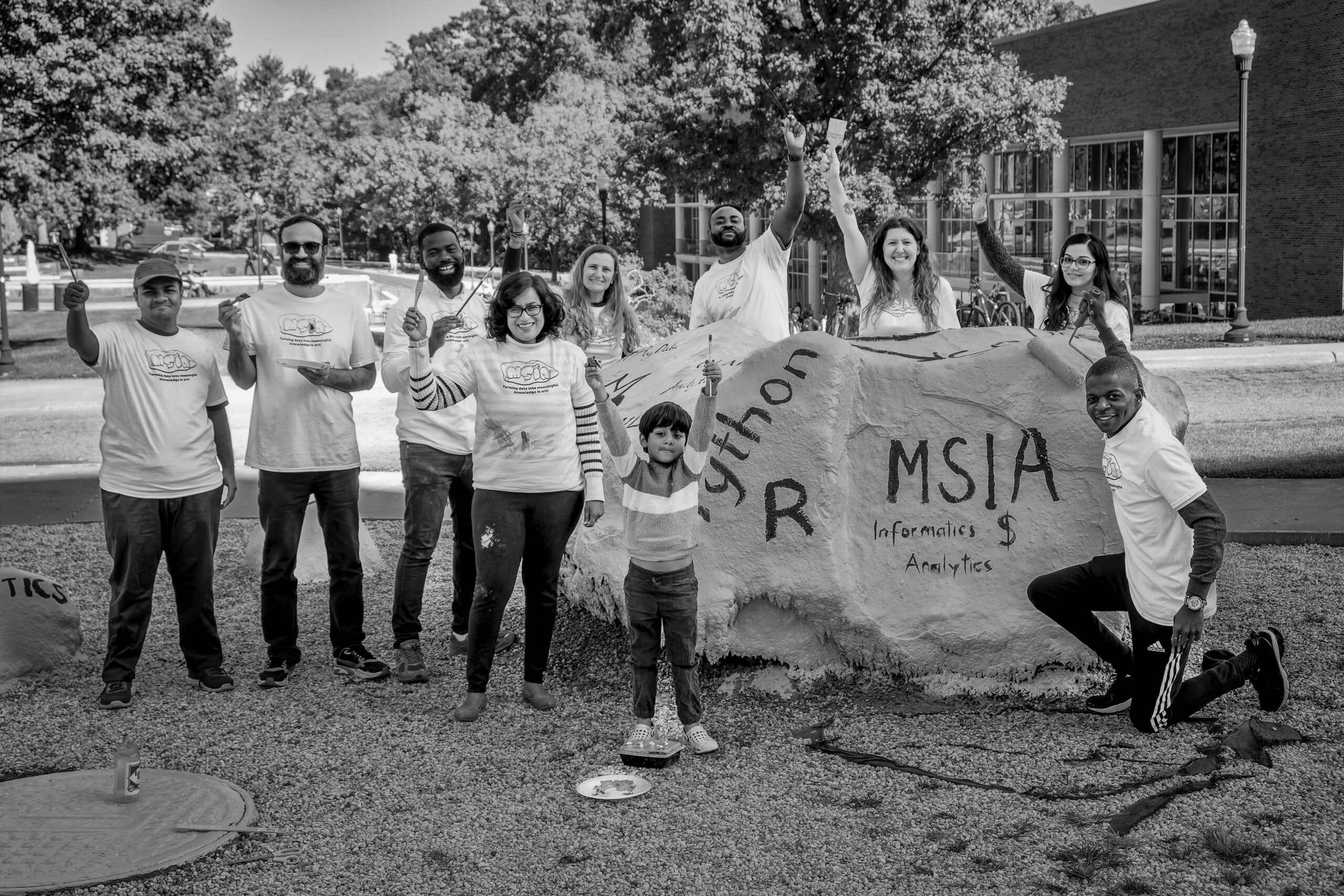 Black and white photo of a group of people standing around a painted rock on the UNCG campus.