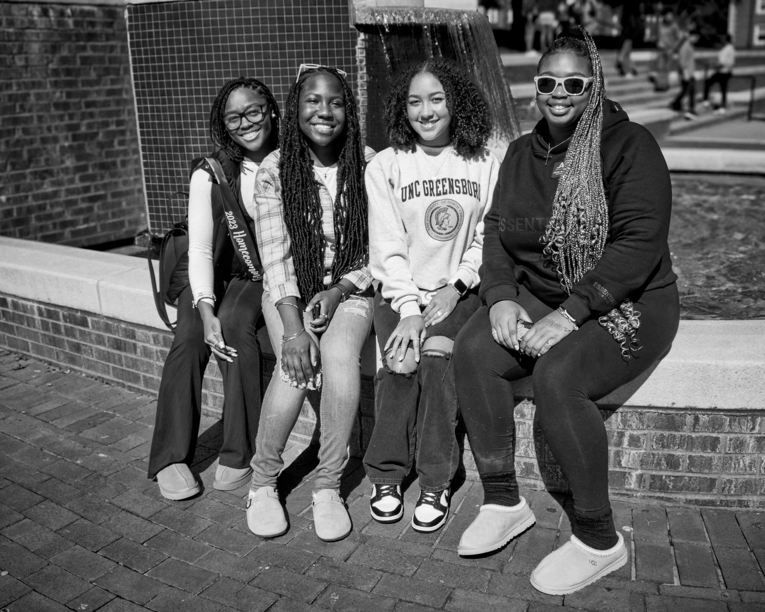 Four smiling UNCG students sit on the edge of the fountain at UNCG William E. Moran Commons and Plaza.