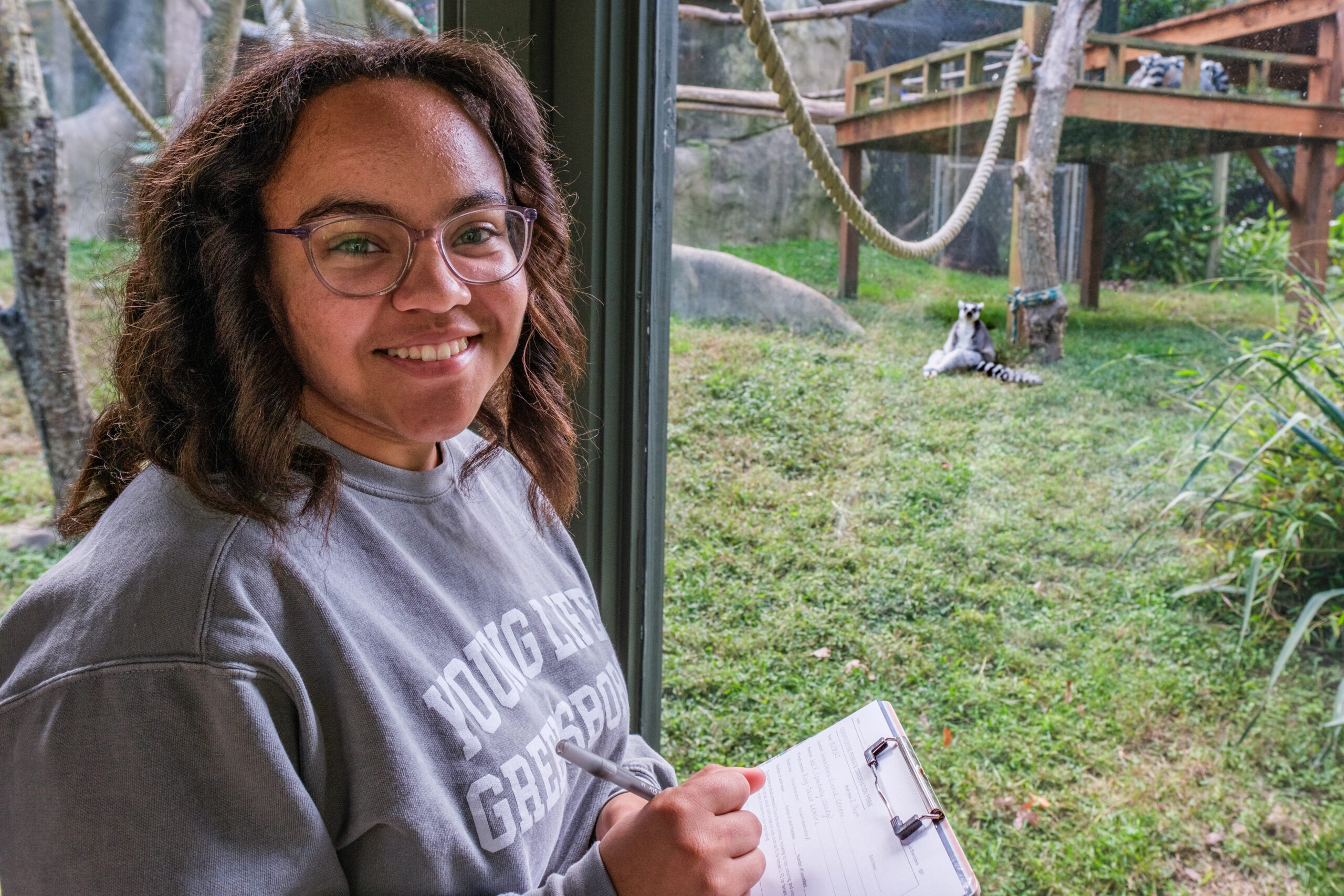 Student smiles holding clipboard in front of a glass-enclosure at the zoo. Behind the glass is a lemur looking towards her.