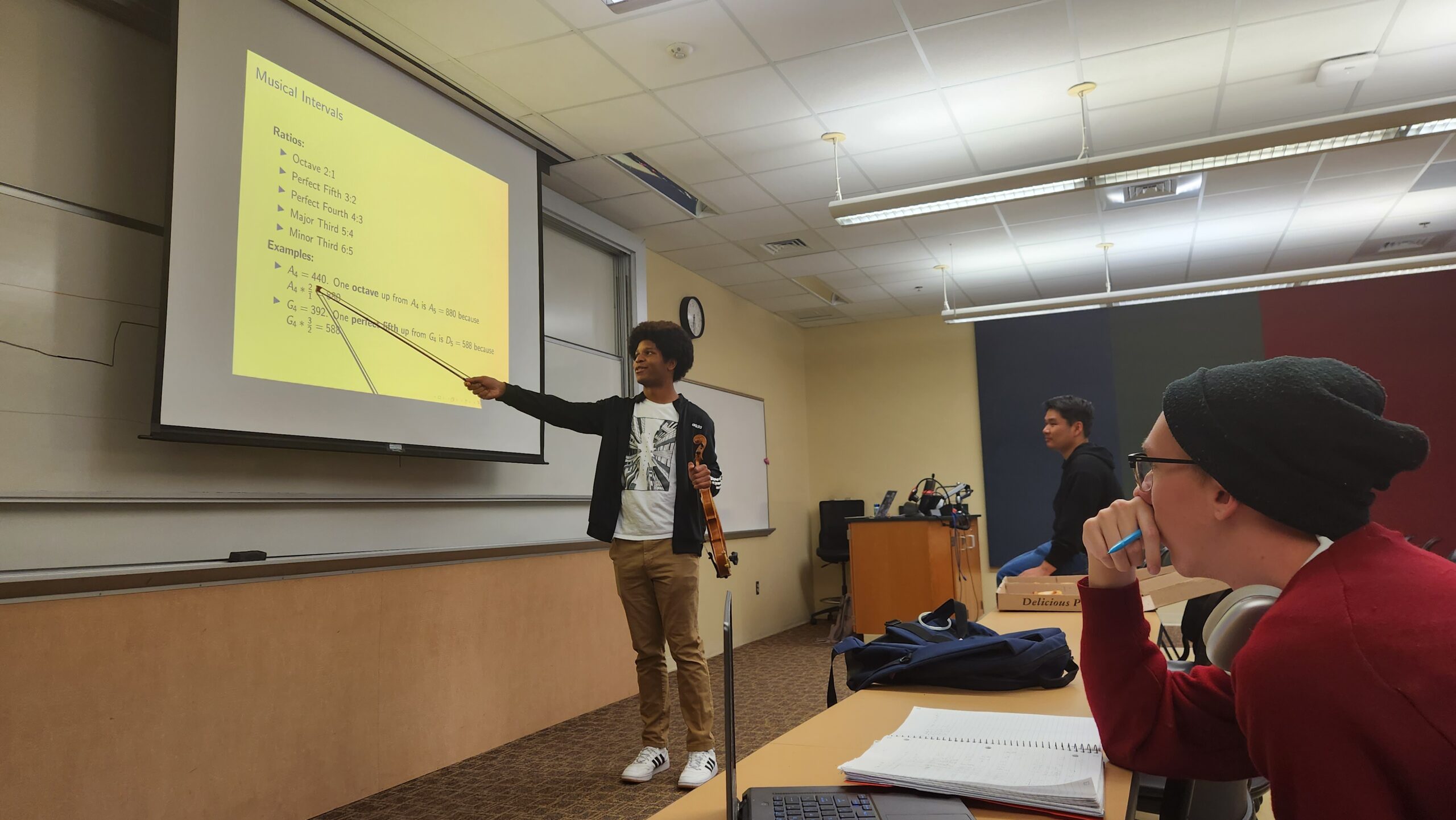Student stands in front of a projector screen, holding a violin in one hand. In the other hand, he points at a presentation with a violin bow.
