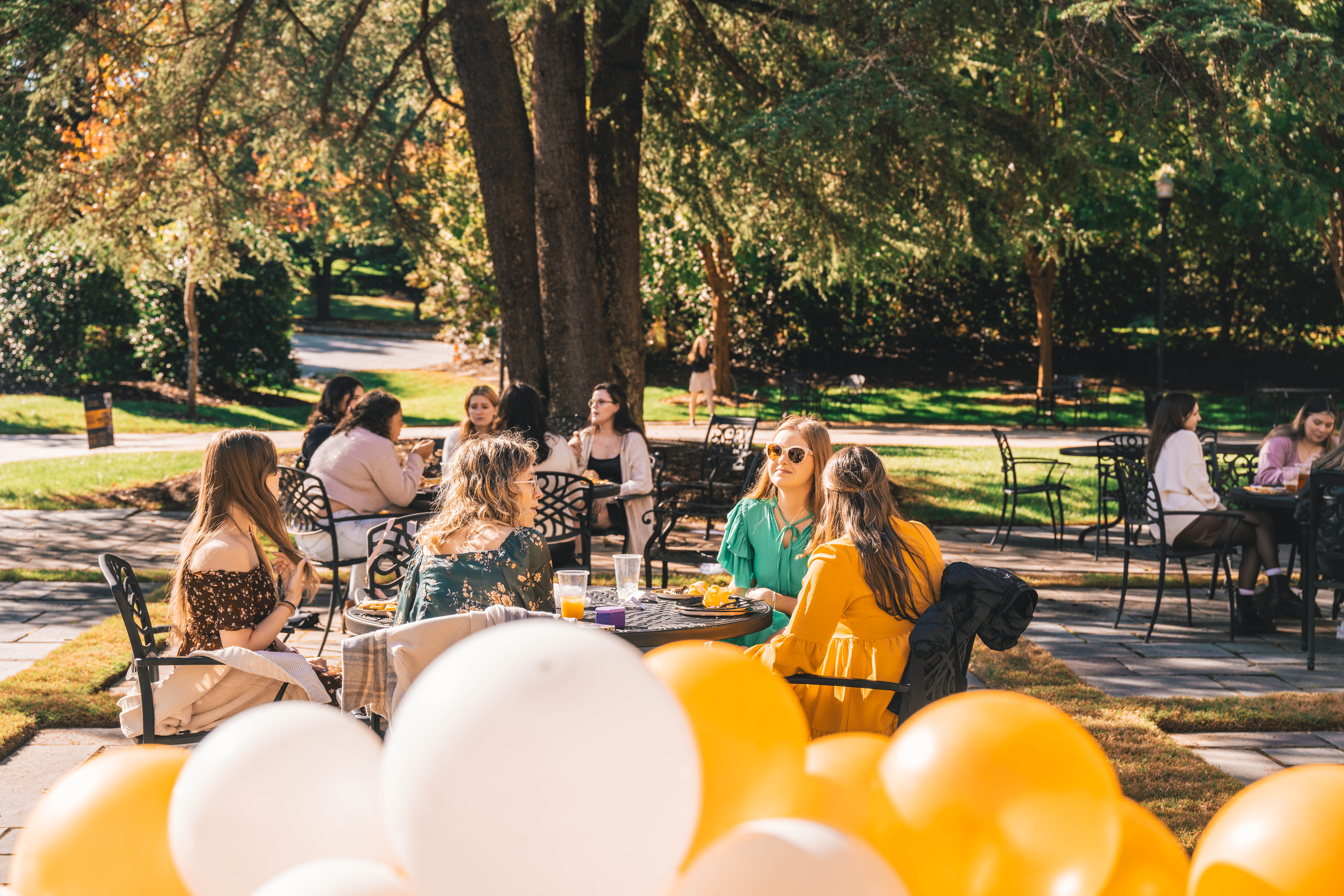A group of women is seated at outdoor tables with fall trees in the background and white and yellow balloons in the foreground.