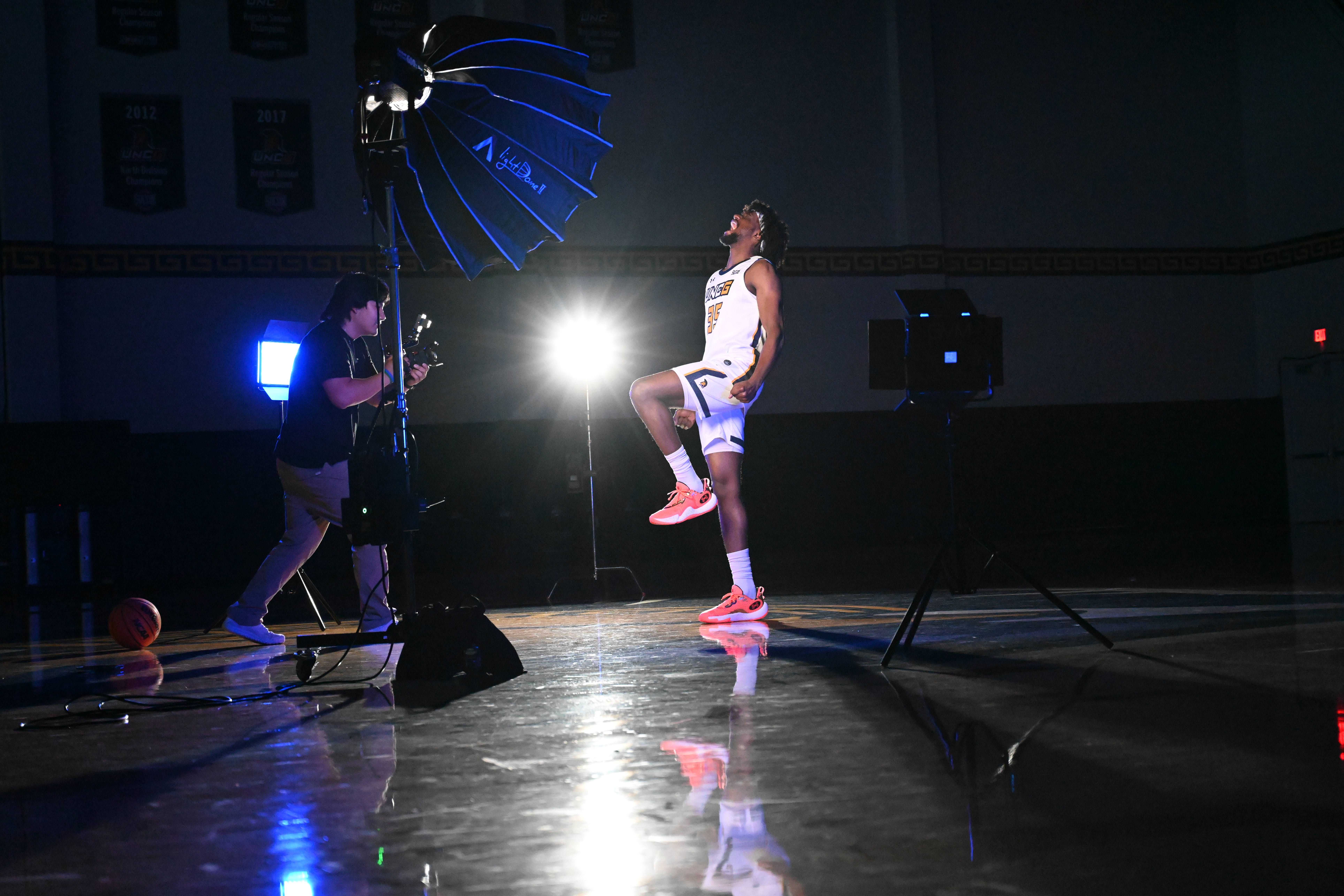 A media studies student stands behind a large camera taking video of basketball player who poses for the team's intro video.