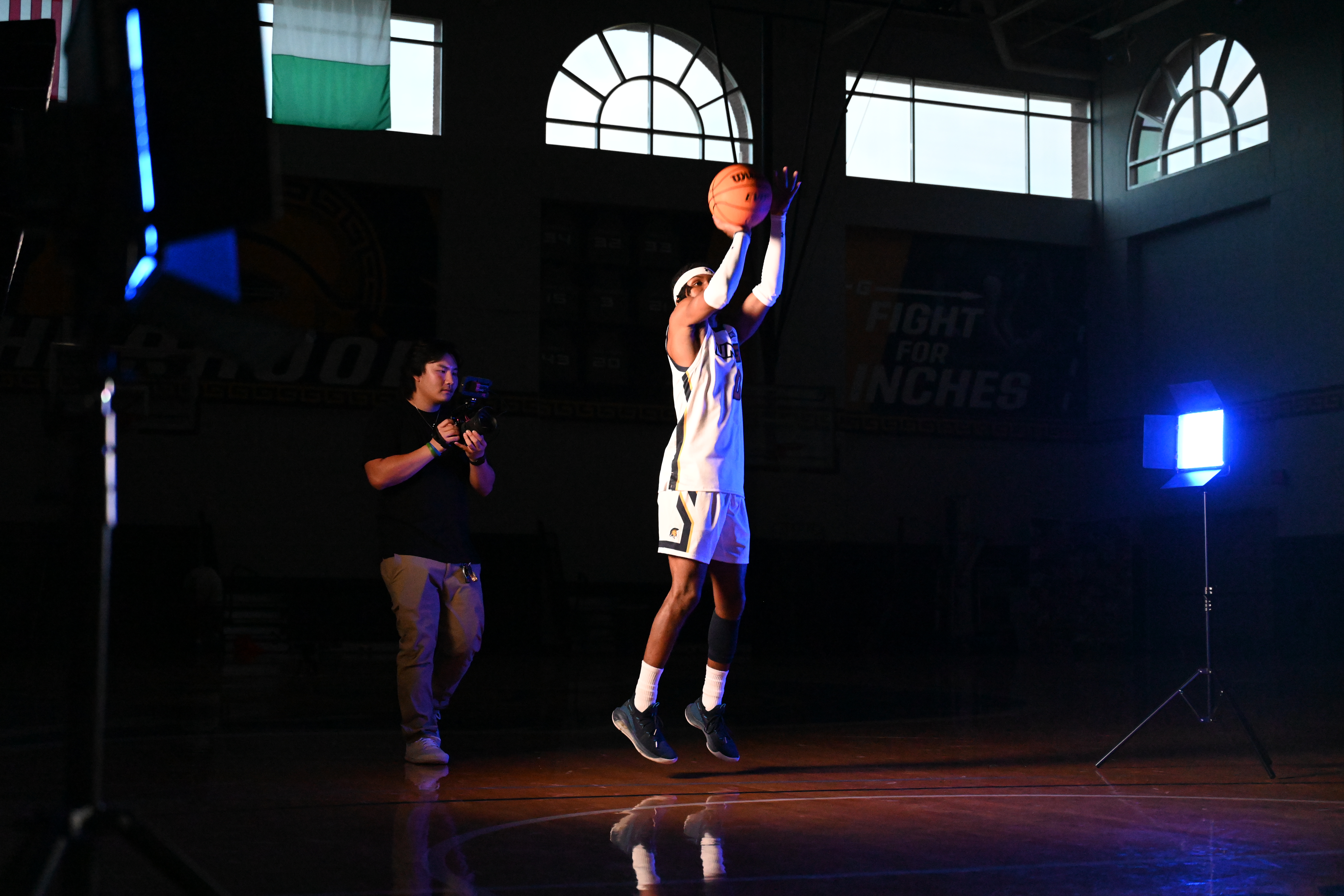 A media studies student stands behind a large camera taking video of basketball player who poses with a basketball for the team's intro video.