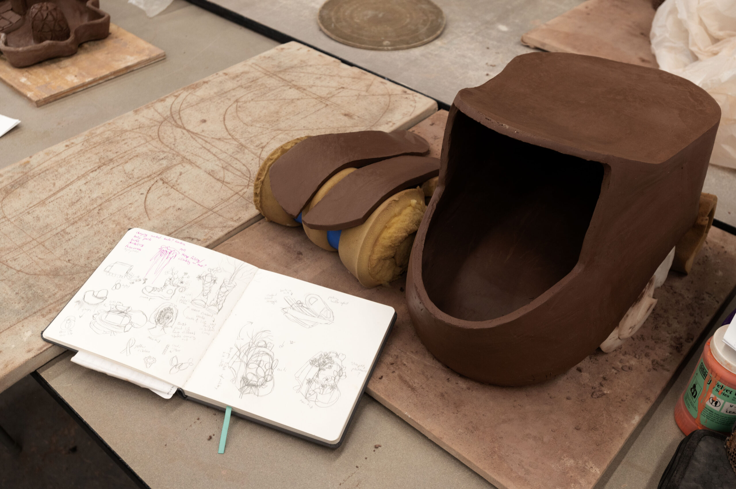 A sketchbook sits next to a half-finished ceramic scultpure of a backpack.