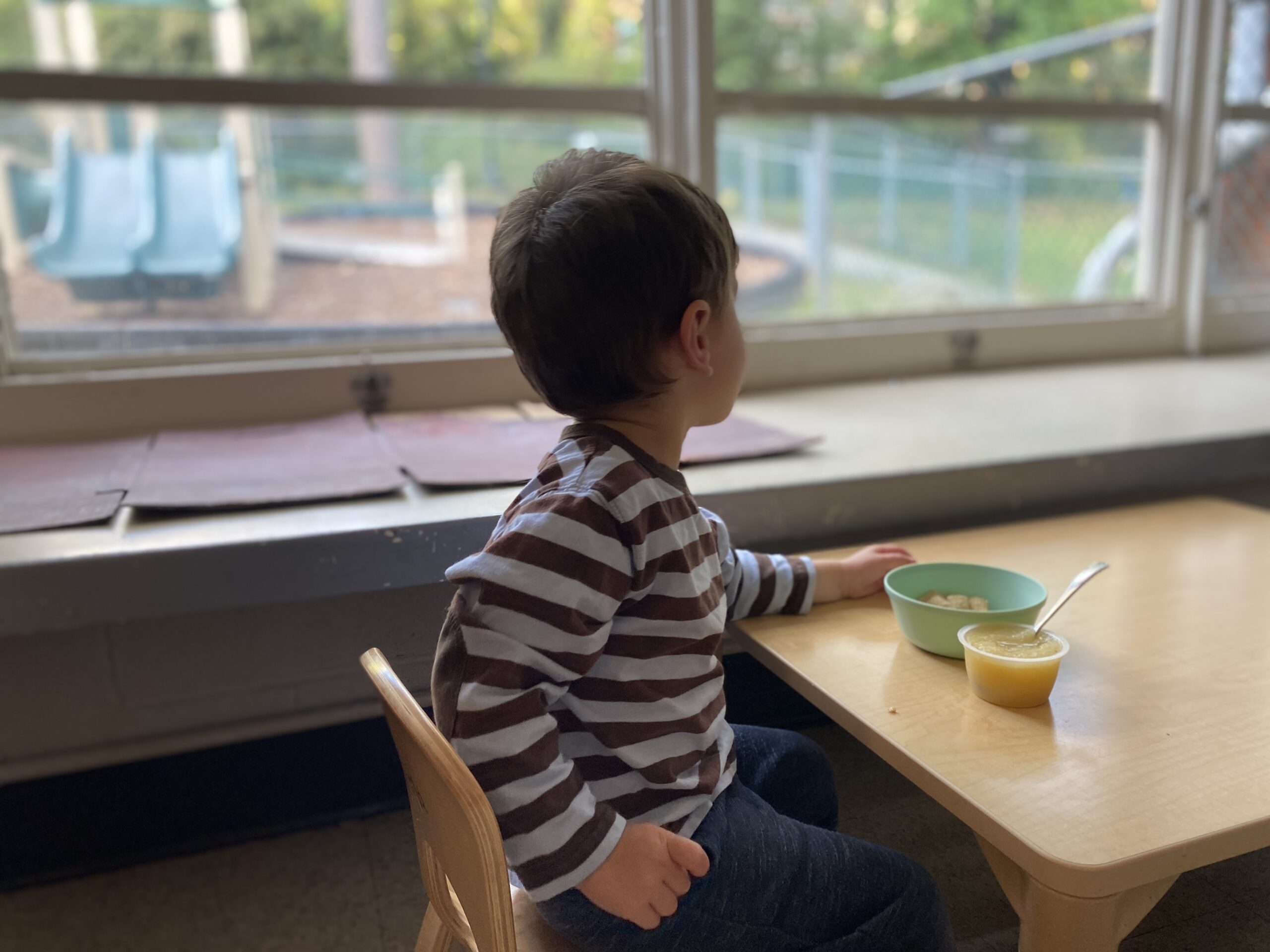 A boy eats at his desk and looks out a window.