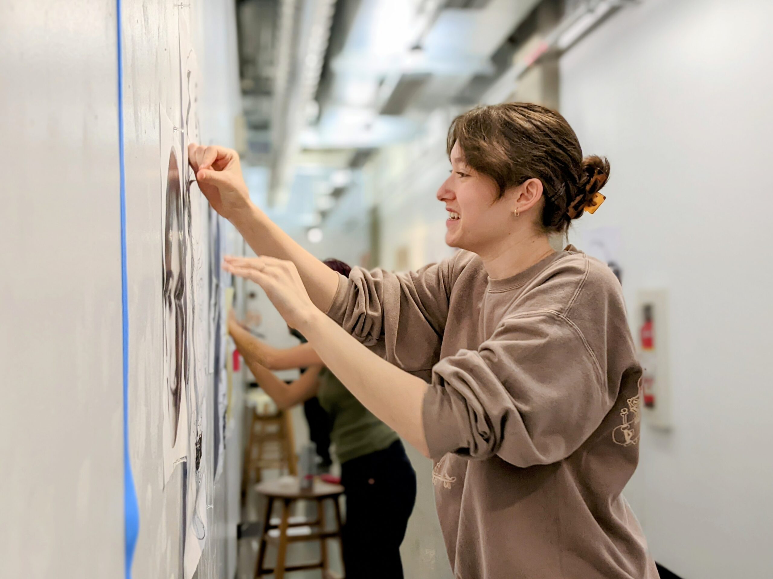 Student tacks art on a white hallway wall.