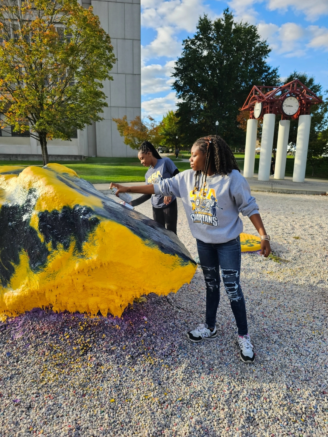 Two students paint a rock blue and yellow on a college campus with a clock tower in the background.