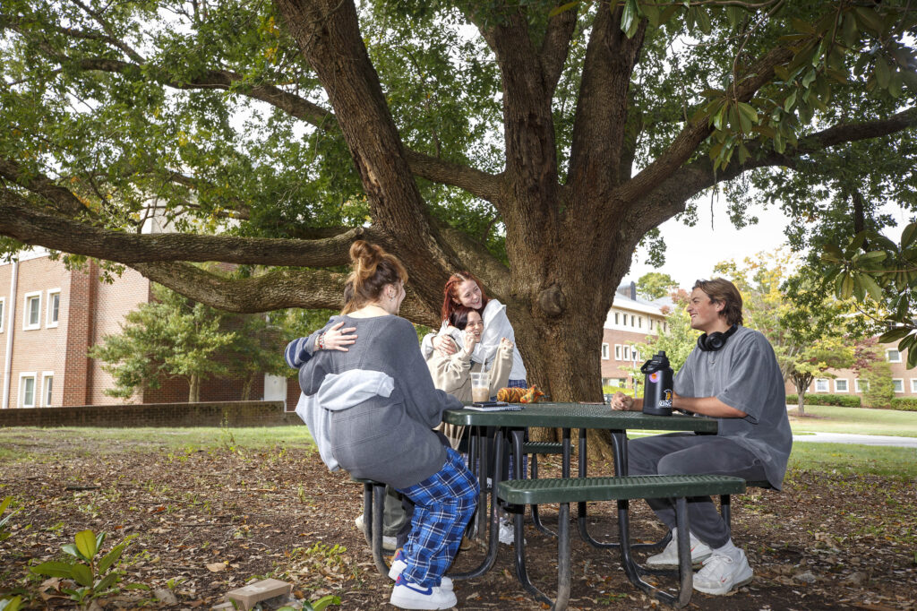 A group of five students, with two pairs embracing each other, sit around a picnic table on the UNCG campus. A large tree with sprawling branches is in the background.