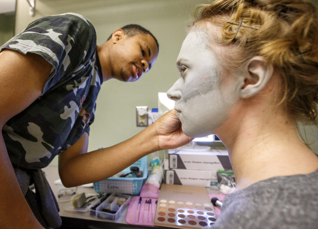 A student applies grey theatre makeup to the face of another student in the Brown Building at UNCG.