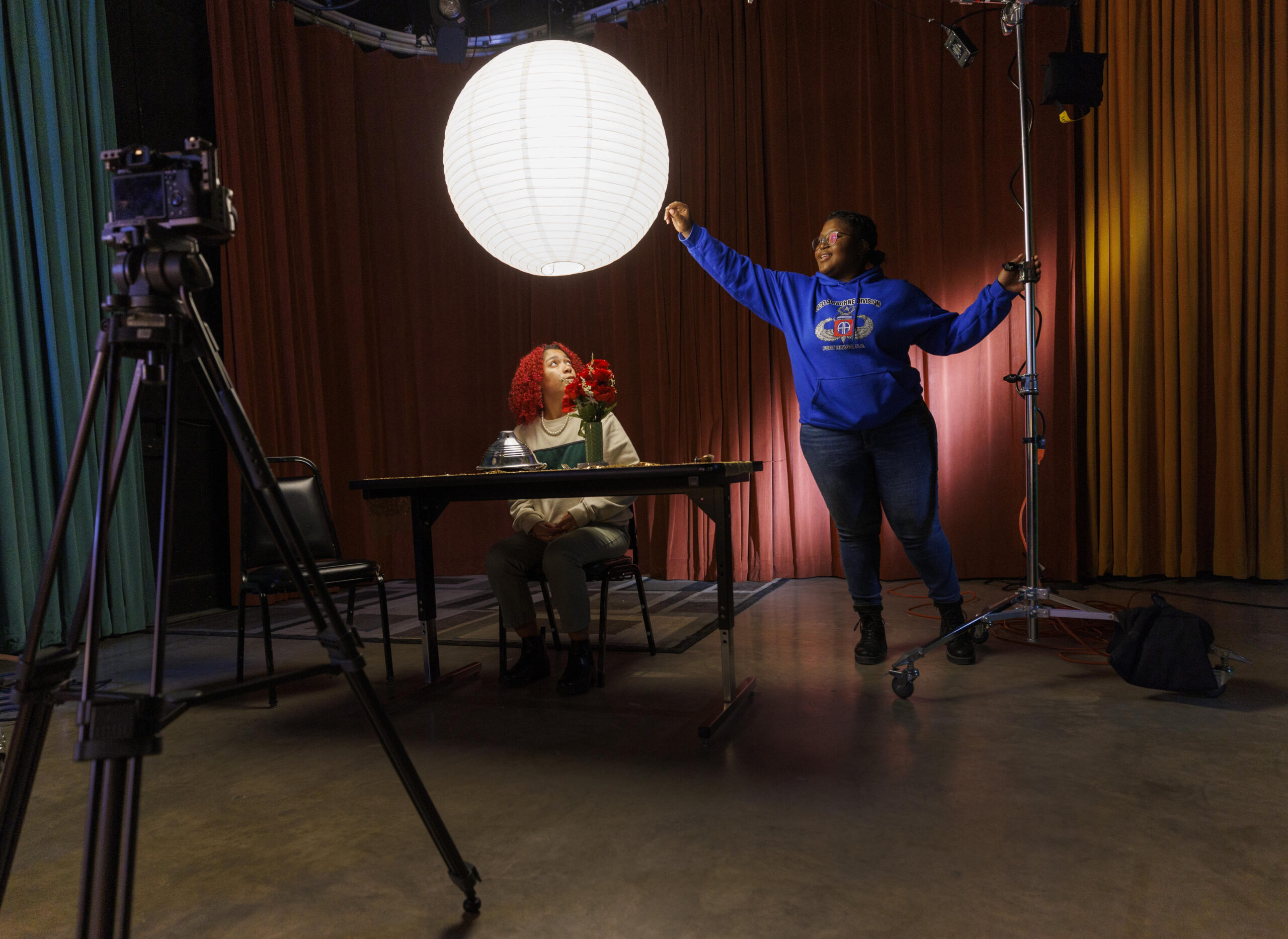 A student is seated at a table in a theater with a camera pointed at her. Another student adjusts lights for her.