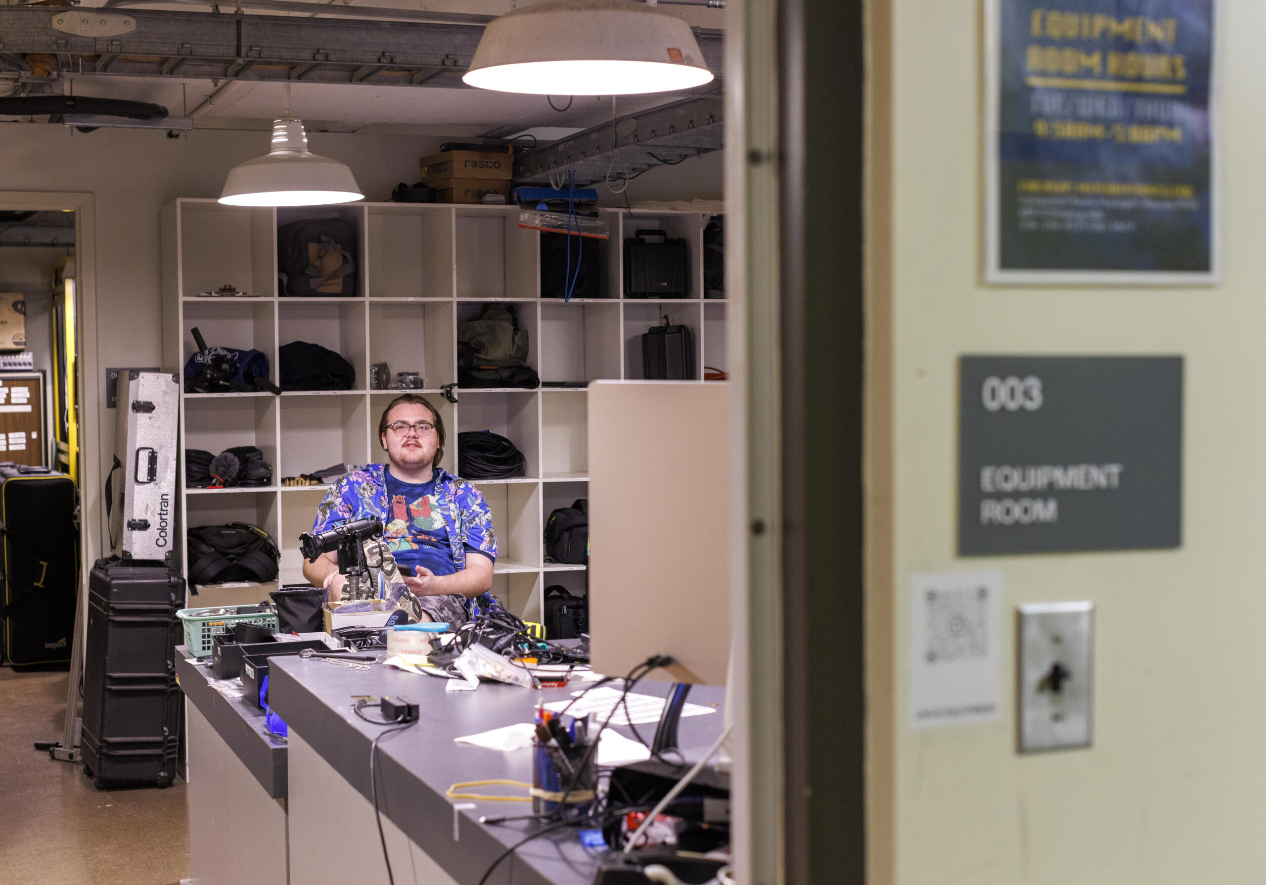 A UNCG student sits in front of shelves of filming equipment.