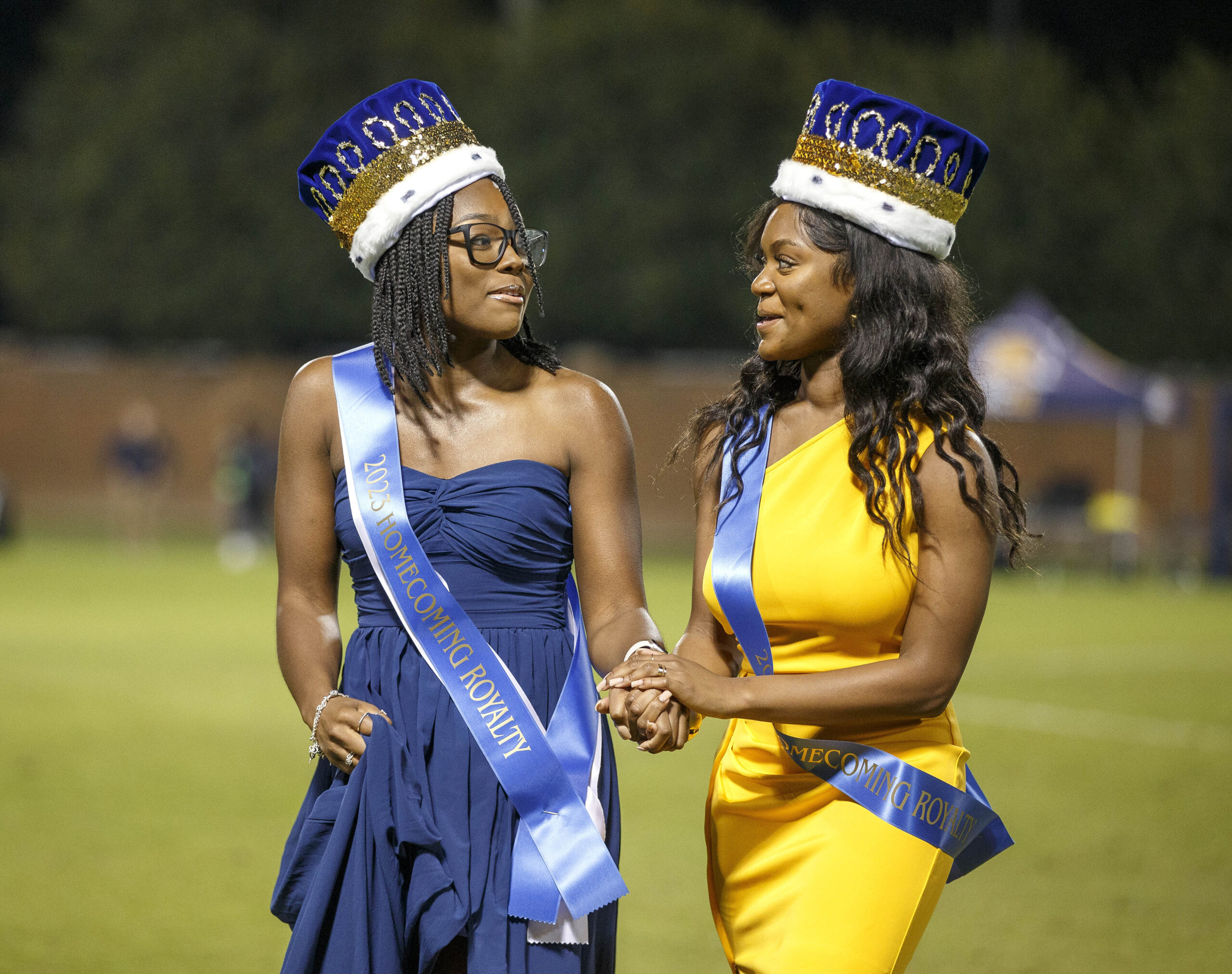 A woman in blue dress holds hands with a woman in a yellow dress as the homecoming royalty are named.