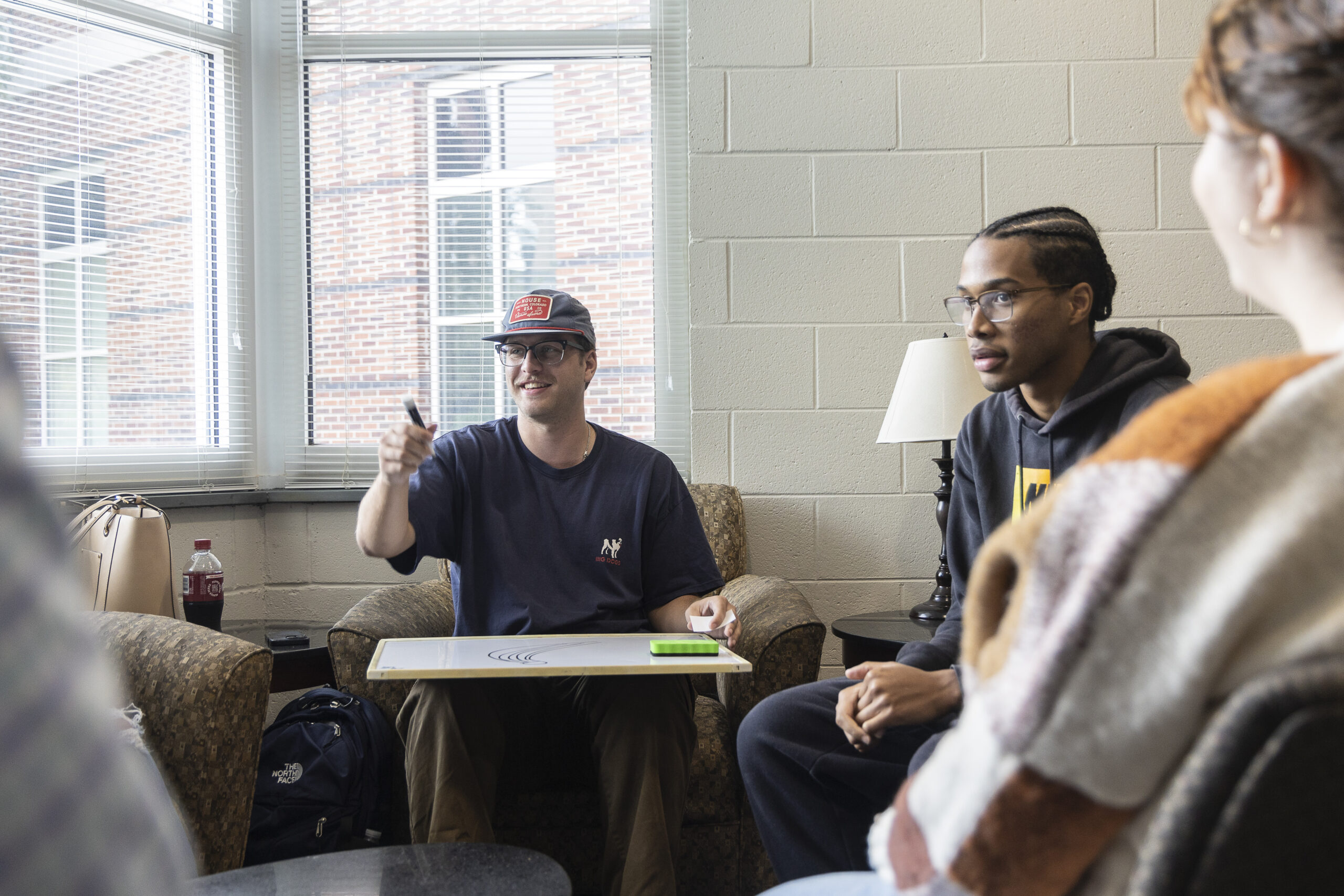 Two students are seated together, one wearing a cap and gesturing toward something, while the other, wearing glasses, listens attentively.