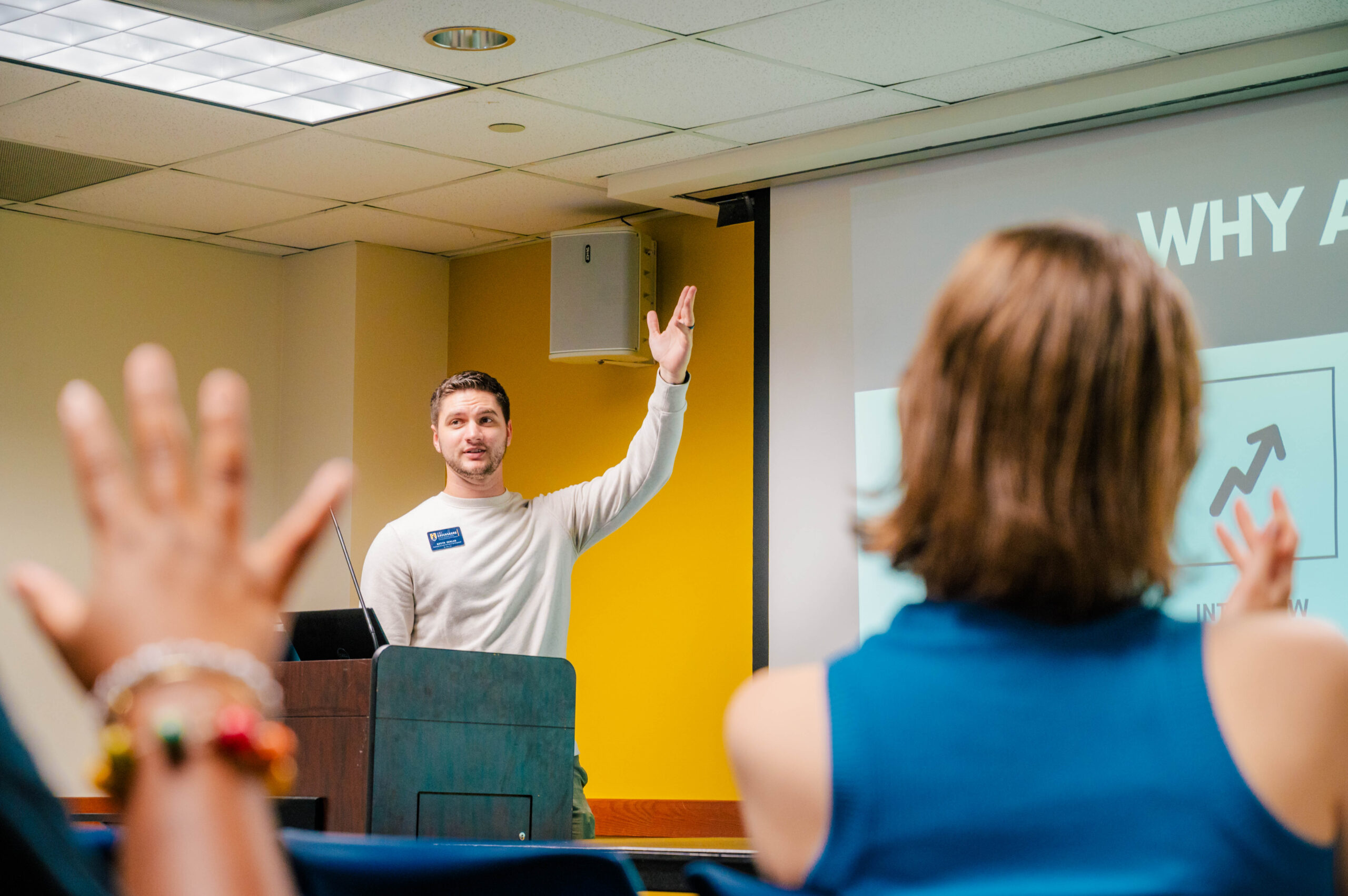 Presenter stands behind a podium in front of a class of students who are raising their hands.