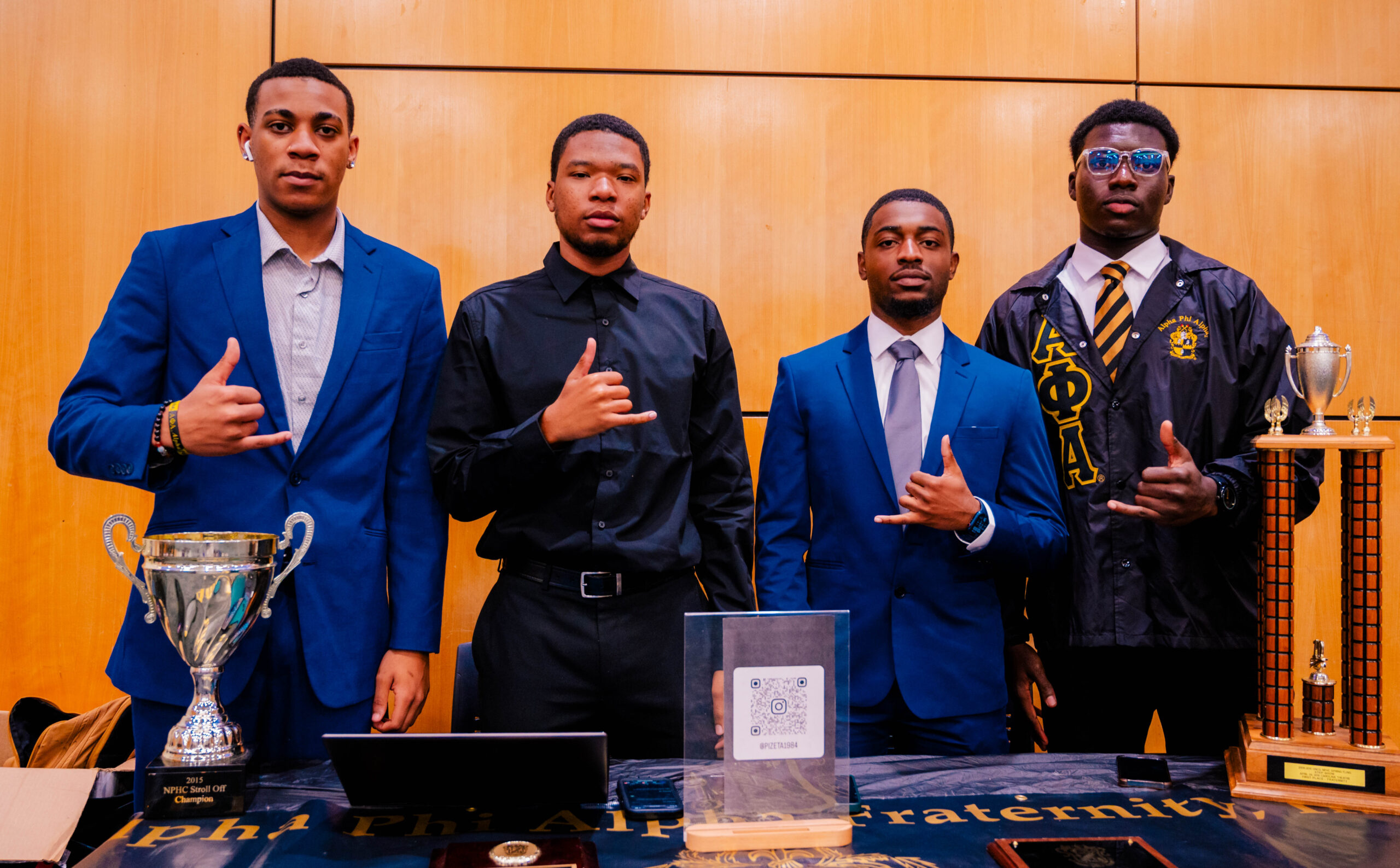 Four students wearing black and royal blue outfits make a hand gesture with their thumbs and pinky fingers extended. They are members of Alpha Phi Alpha fraternity stand in front of a table with fraternity paraphernalia in the Elliott University Center.