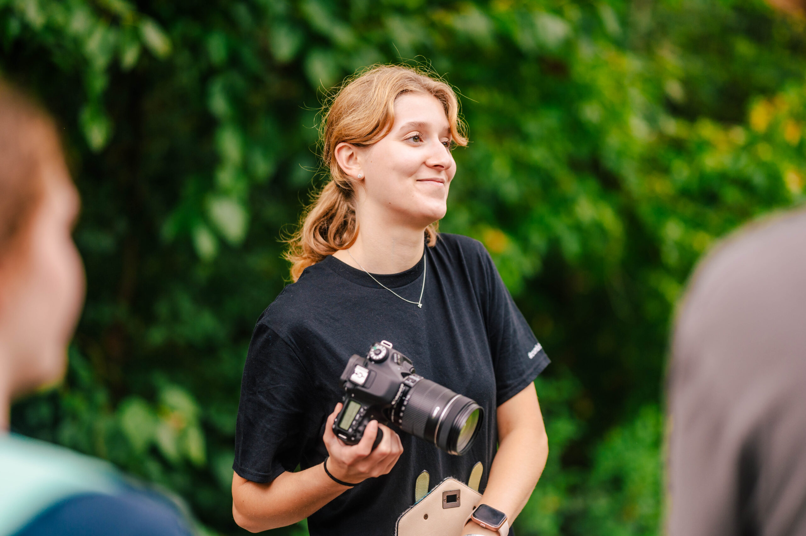 A person with shoulder length red hair gathered in a ponytail holds a digital camera as they stand outside on College Avenue.
