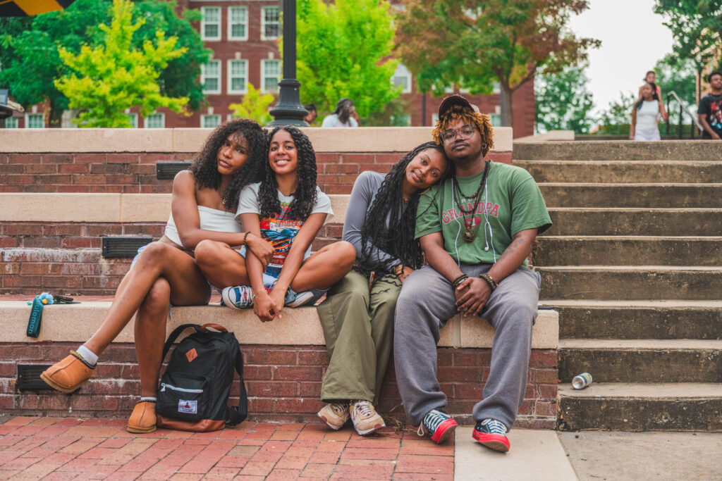 Four students of color, seated on brick steps at the Fountain outside of Moran Commons, look straight ahead. One student embraces another while a third student lays her head on the shoulder of a fourth.
