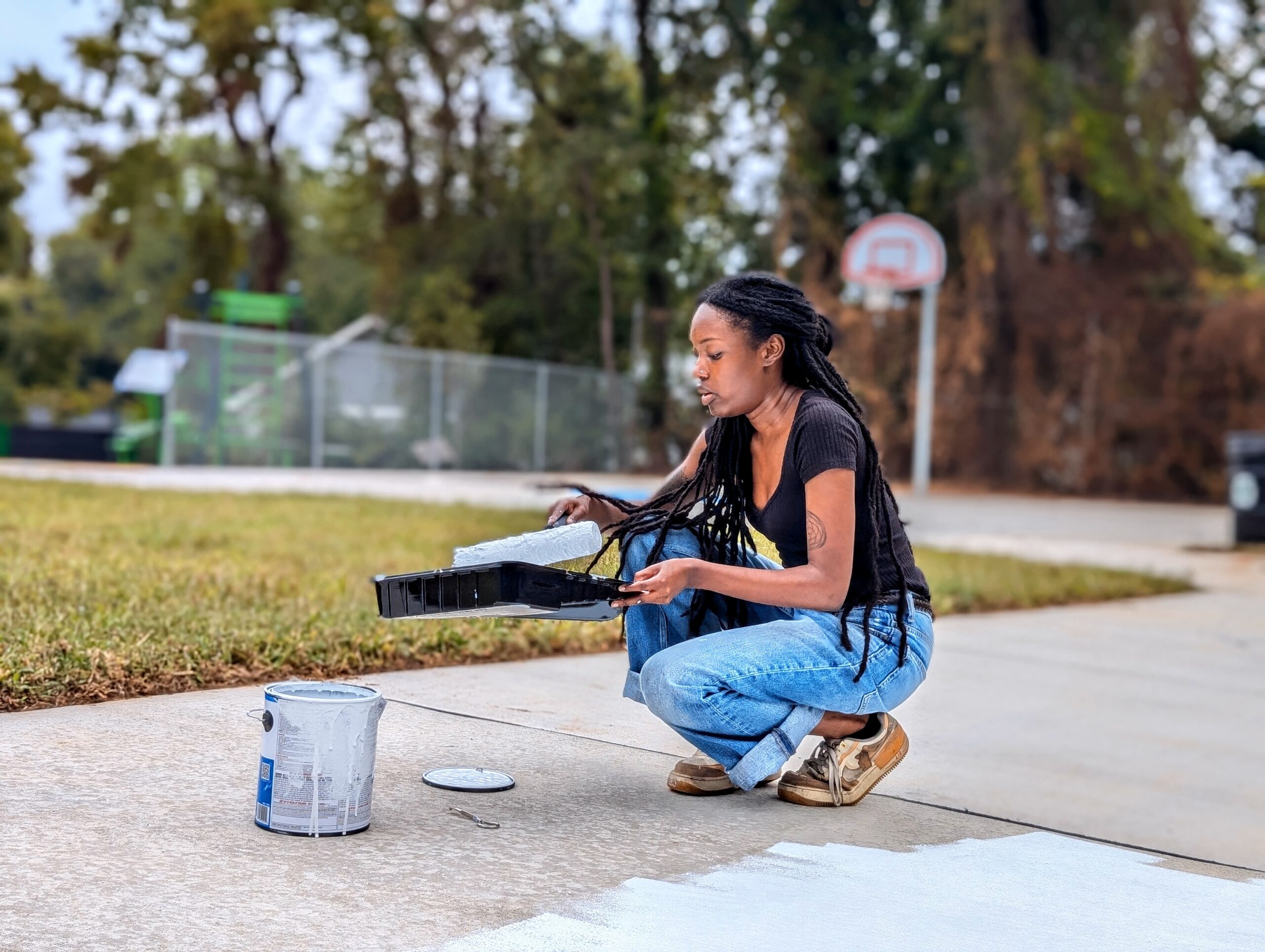 Student paints a sidewalk in a part with a basketball court behind her.