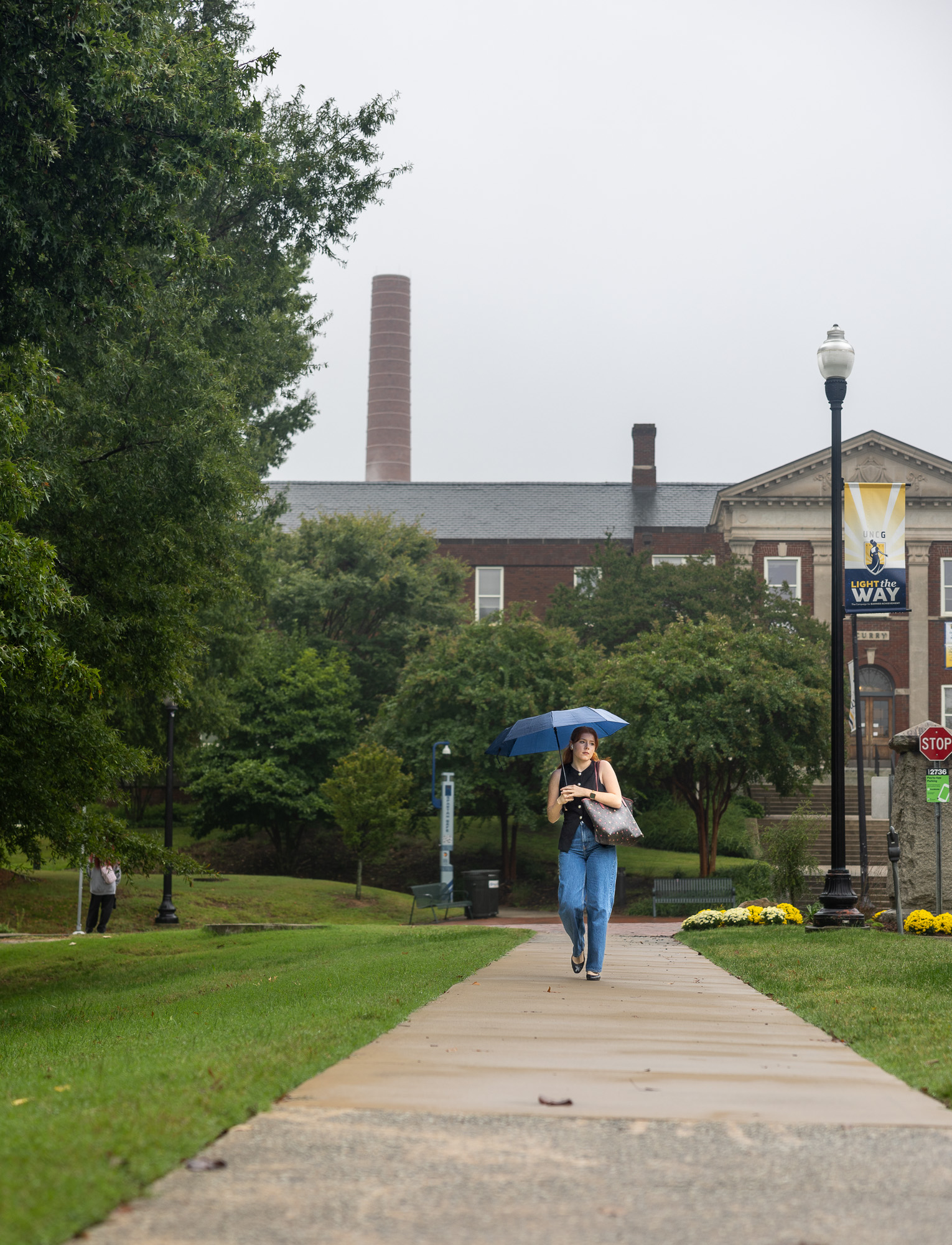 A girl holding a blue umbrella, walking on the sidewalk of college campus.