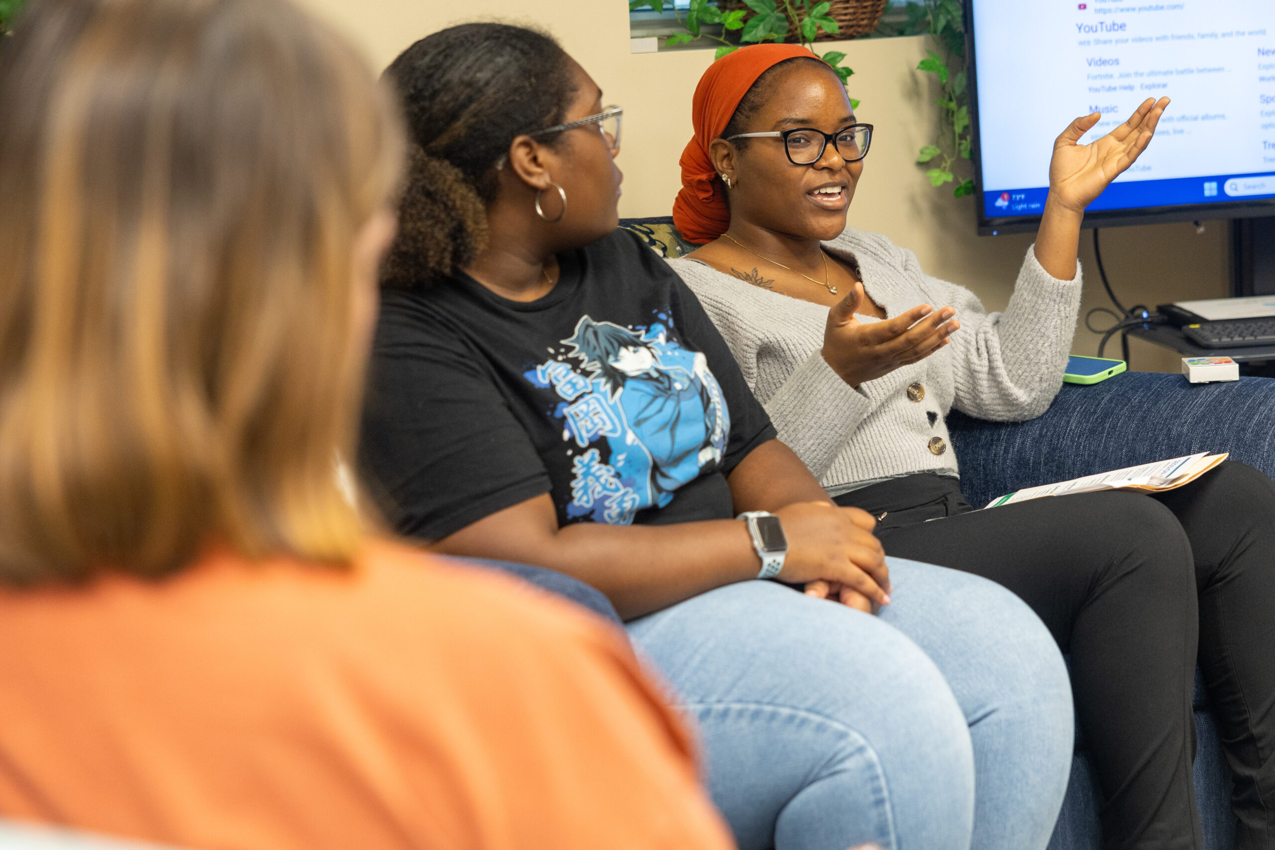 Three women are seated in room with a digital screen hanging on a wall behind them. A woman wearing a dark orange head scarf speaks, her hands raised for emphasis.