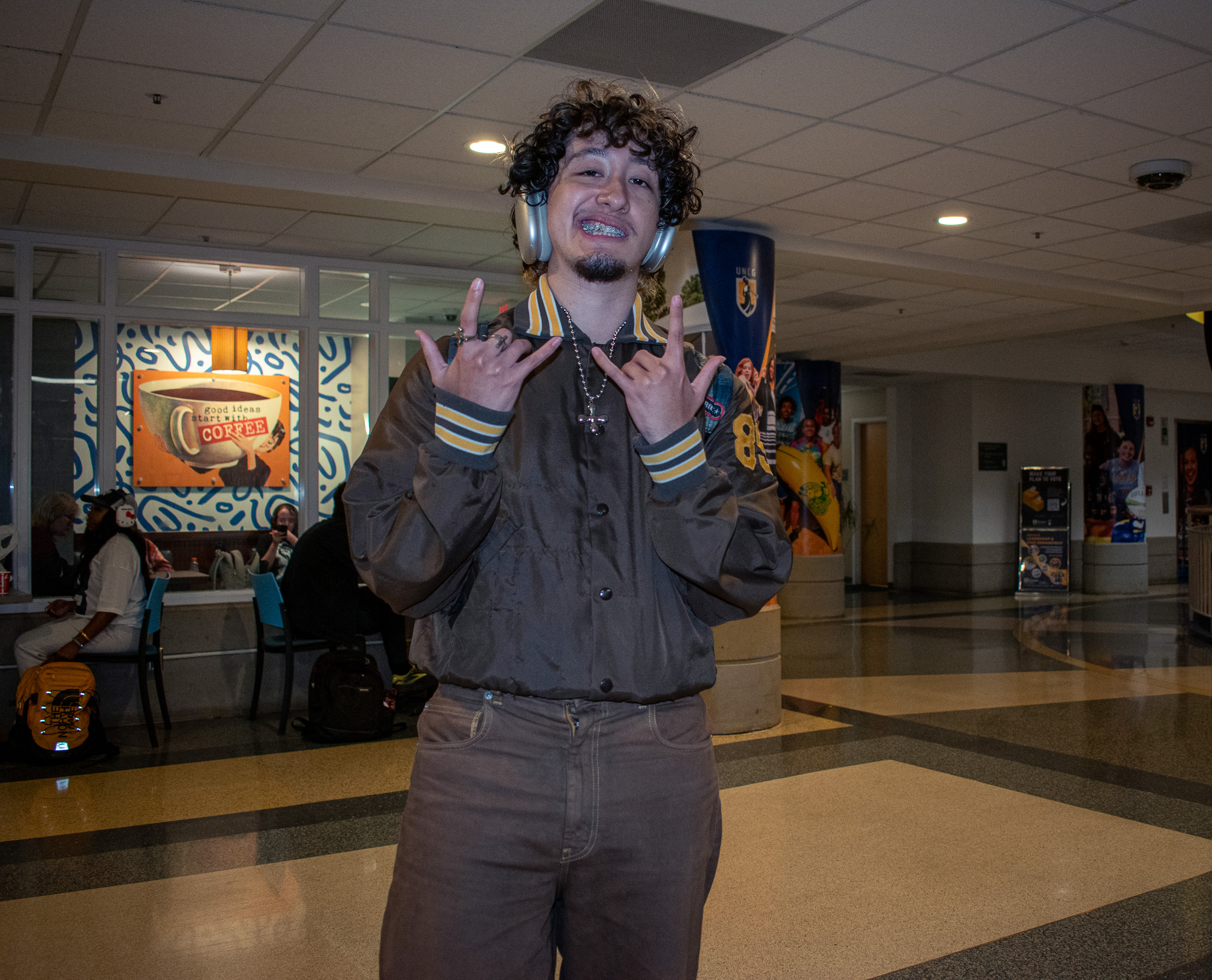 Student in headphones flashes "I love you" hand signs and grins for the camera in a hall in EUC.