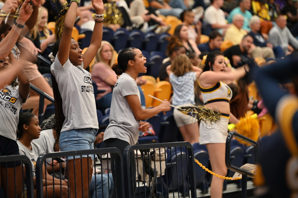 Two women, facing right and wearing shirts that read "UNCG student athlete," stand up and cheer during a volleyball game in Fleming Gym. Surrounding them are a UNCG cheerleader and other spectators at the game.