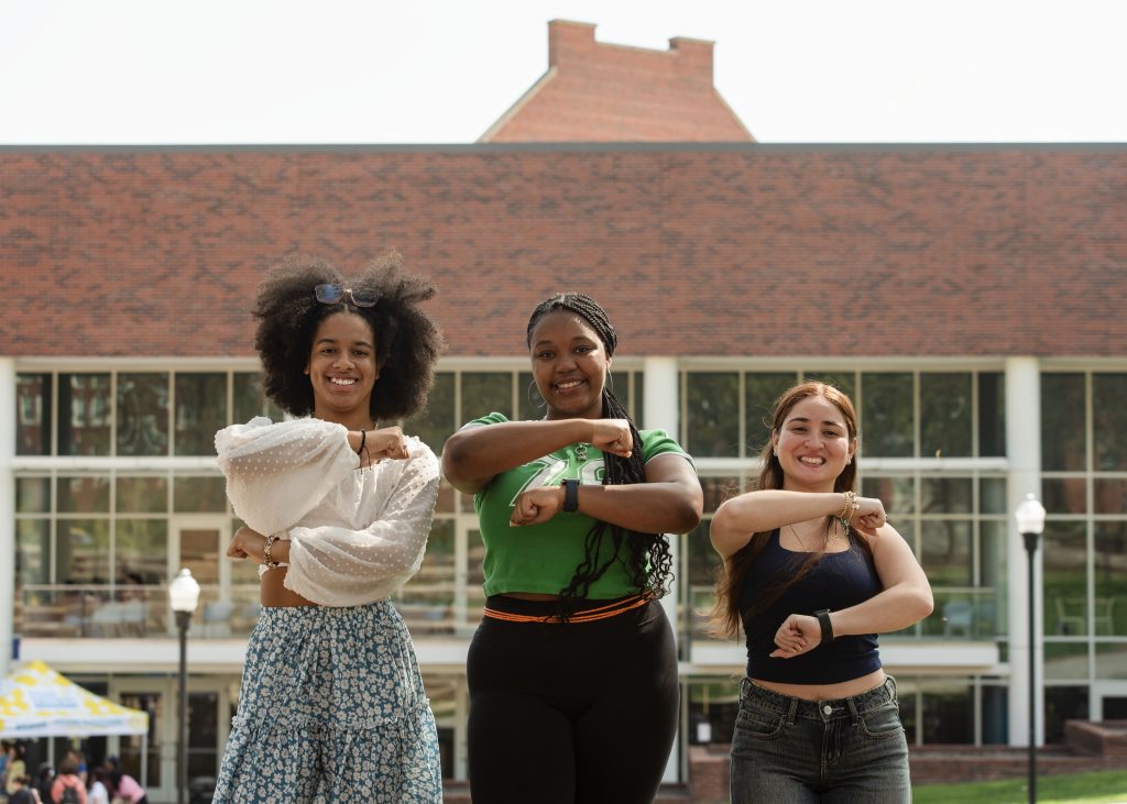 Three girls stand in front of Moran Commons and make the G sign with their arms.