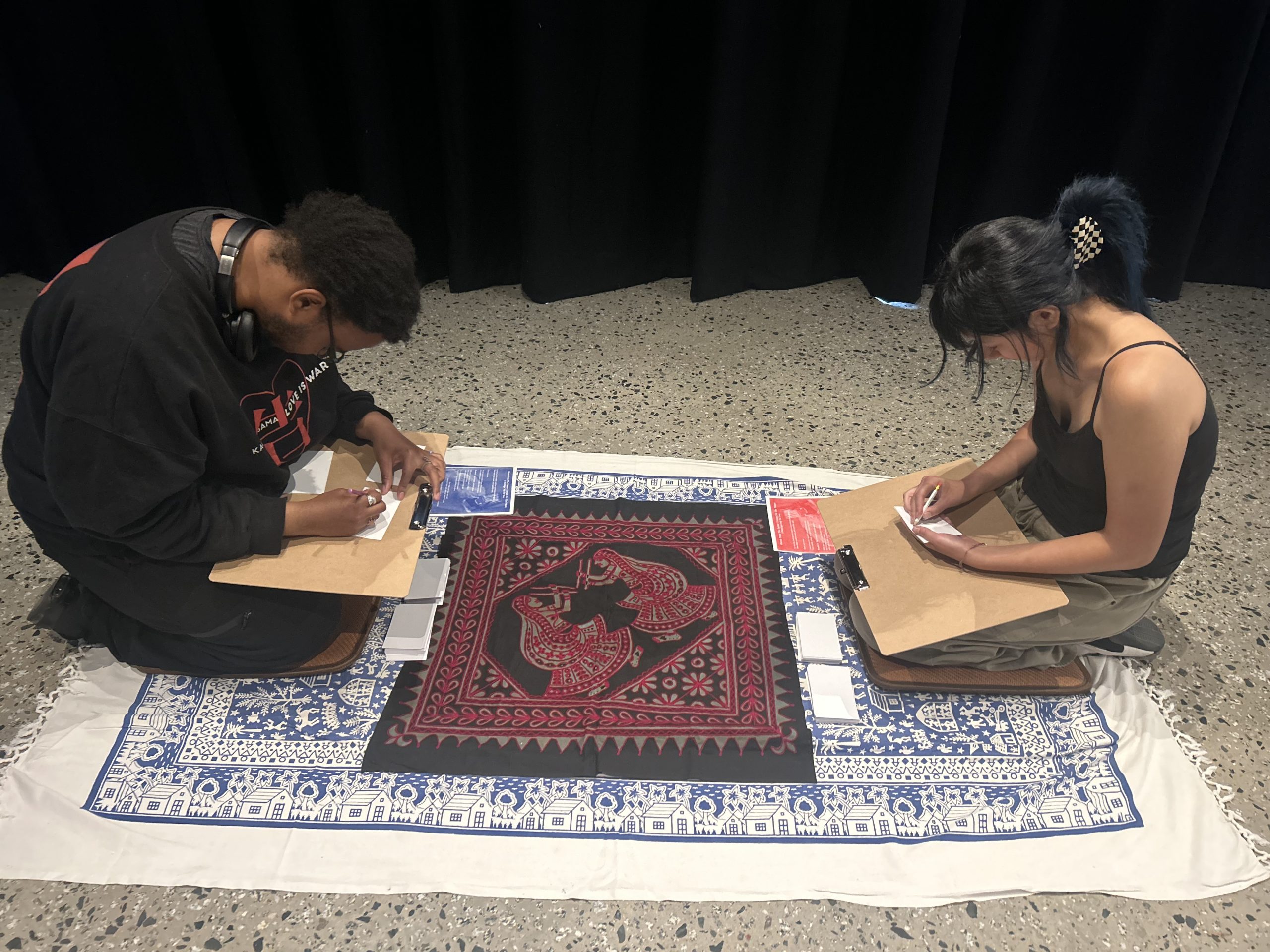 2 students sit on either side of a game board on a rug on the floor and make notes in notebooks.