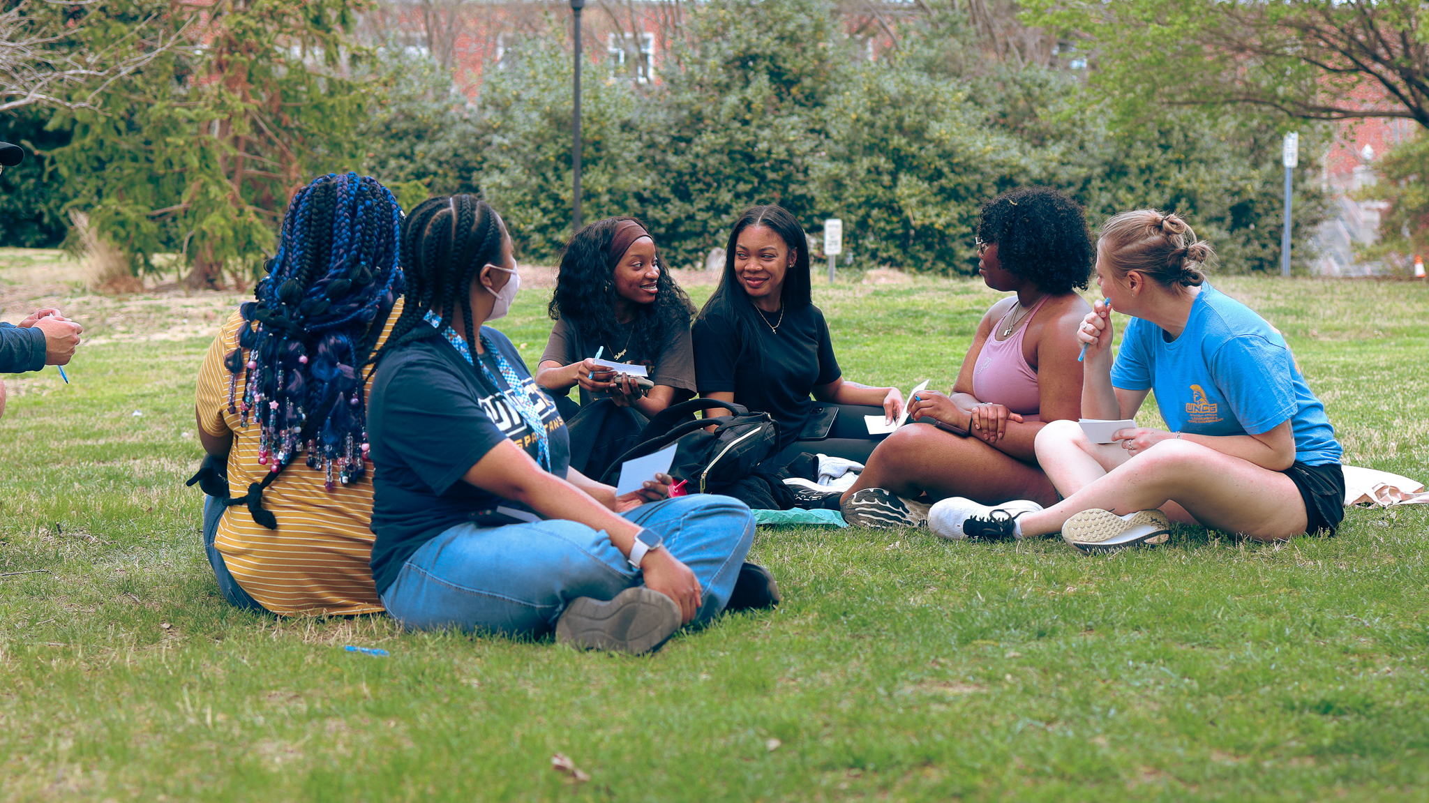 A group of women holding index cards and talking sit on a grass lawn at Foust Park.
