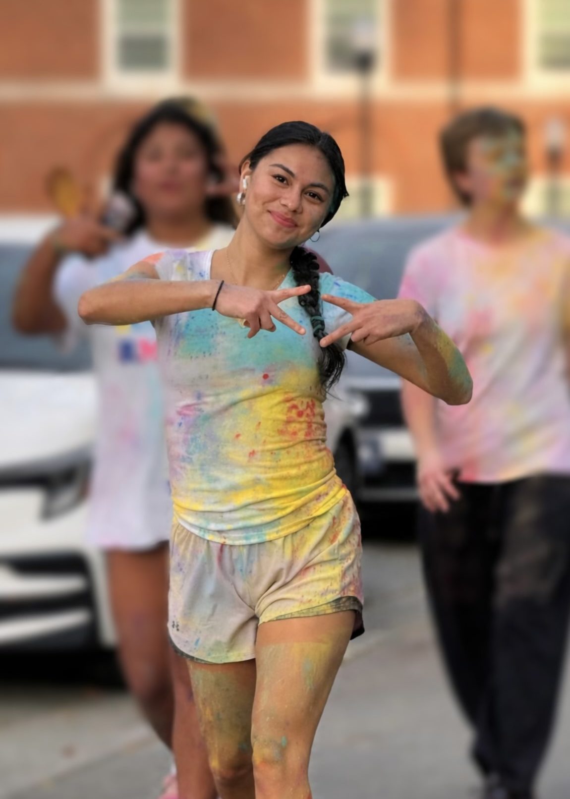 Girl makes a Greek sign with her hands and smiles as she walks with others who are dressed in t-shirts with paint splotches.