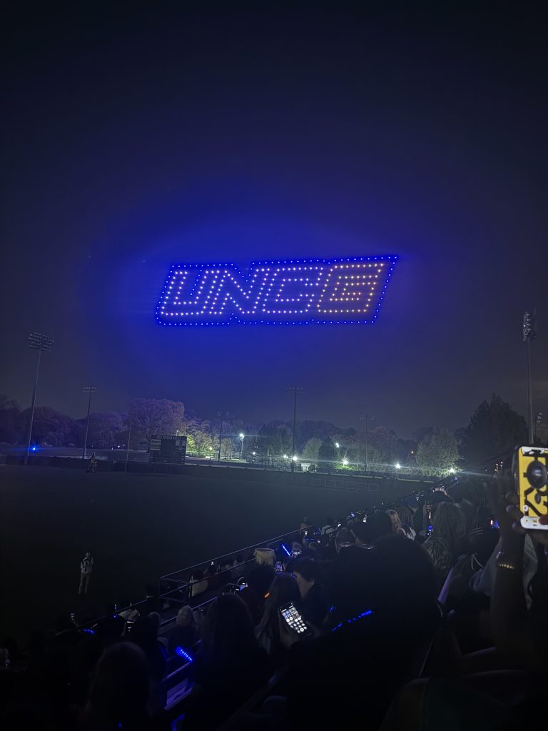 A drone show displays the UNCG logo in blue and gold lights in the night sky above the UNCG soccer stadium.