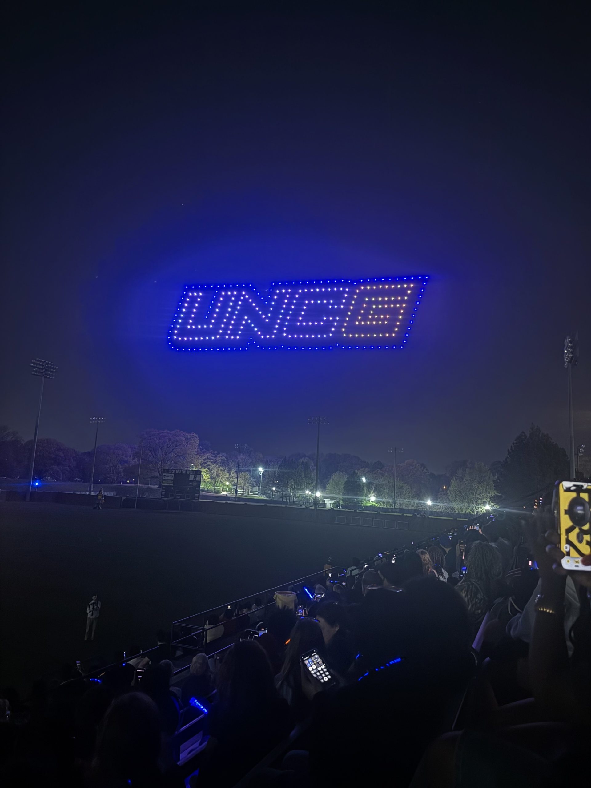 A drone show displays the UNCG logo in blue and gold lights in the night sky above the UNCG soccer stadium.