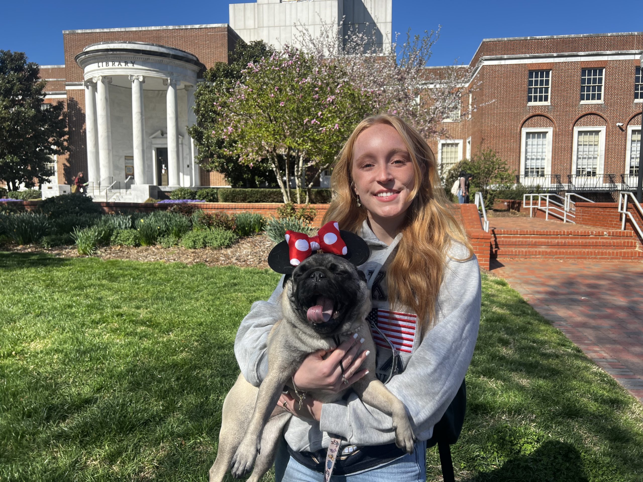 Girl holds a pug with a polka dot bow on its head in front of Jackson Library.