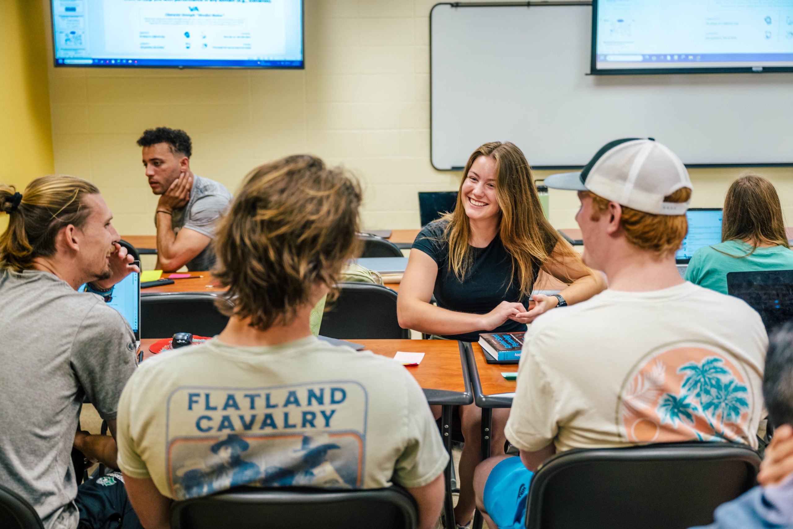 College students engaged in a classroom discussion with laptops and notebooks on desks in front of a whiteboard and projection screens.