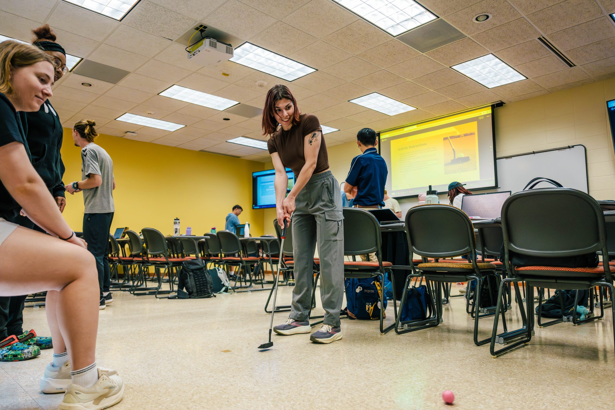 A person swings a golf club in a classroom setting with chairs and a projector screen in the background.